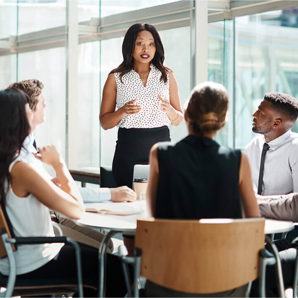 A woman with a Ph.D. stands and speaks to four colleagues seated around a table in a glass-walled office during a meeting.
