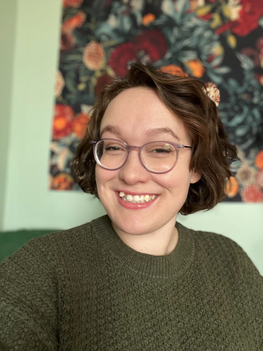 Une personne souriante aux cheveux bouclés et aux lunettes porte un pull-over vert. Une tapisserie florale colorée est suspendue à l'arrière-plan, créant une atmosphère joyeuse.