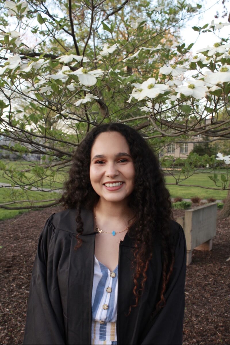 Une jeune femme aux cheveux bouclés sourit chaleureusement, vêtue d'une robe de fin d'études. Elle se tient devant un arbre en fleurs, symbole de bonheur et de réussite.