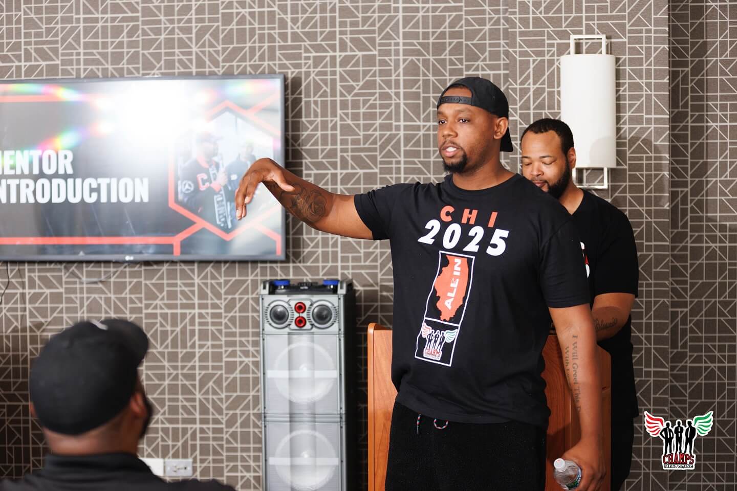 Un homme portant une chemise "CHI 2025", un ancien élève d'Adler, parle de la façon dont le mentorat transforme les vies à un public assis, tandis qu'un autre homme se tient derrière lui sur un podium. Un écran affiche "Présentation du mentor".