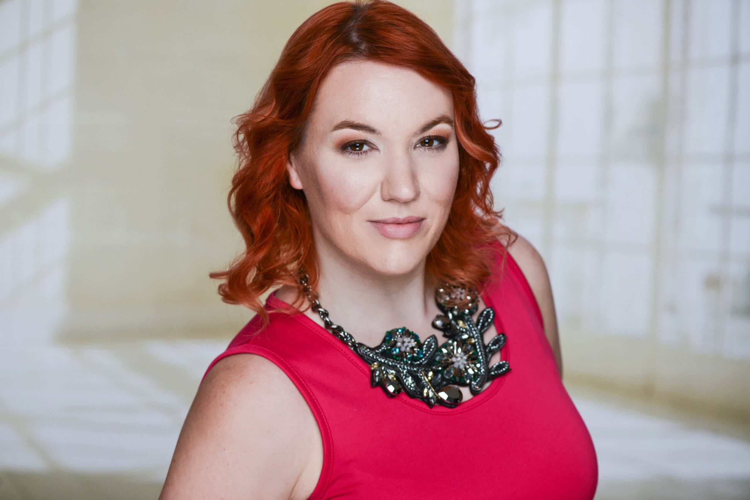 Una mujer pelirroja y ondulada, con un top rosa sin mangas y un gran collar, frente a un fondo suavemente iluminado.