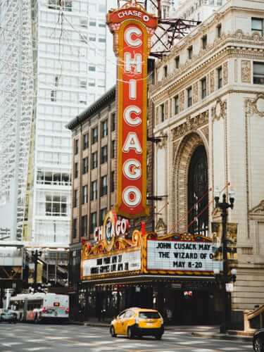 The Chicago Theatre with its iconic marquee sign, located on a city street, hosts a commencement ceremony as a yellow taxi passes by in the foreground.