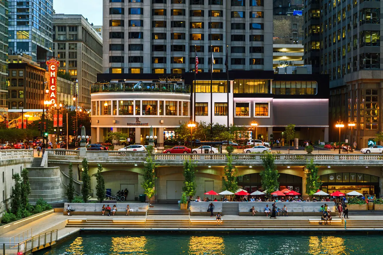 People sit and walk along the Chicago Riverwalk near outdoor cafes, as city buildings and the illuminated Chicago Theatre sign create a lively backdrop, capturing the excitement of commencement season in the city.